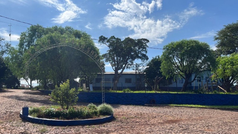 Vê se a rua de pedras, com um canteiro florido no centro, contando com um letreiro com o nome da escola. Ao fundo, o prédio escolar.